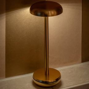 A modern mushroom-shaped table lamp in a brushed gold finish, illuminated and casting a warm light against a textured beige wall.