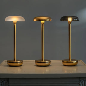 Three modern table lamps with slim gold-finished stems and round bases are displayed on a light-colored console table against a dark gray wall. The left lamp has a frosted white dome shade, the middle lamp has a brushed gold dome shade, and the right lamp has a smoked glass dome shade, all illuminated.