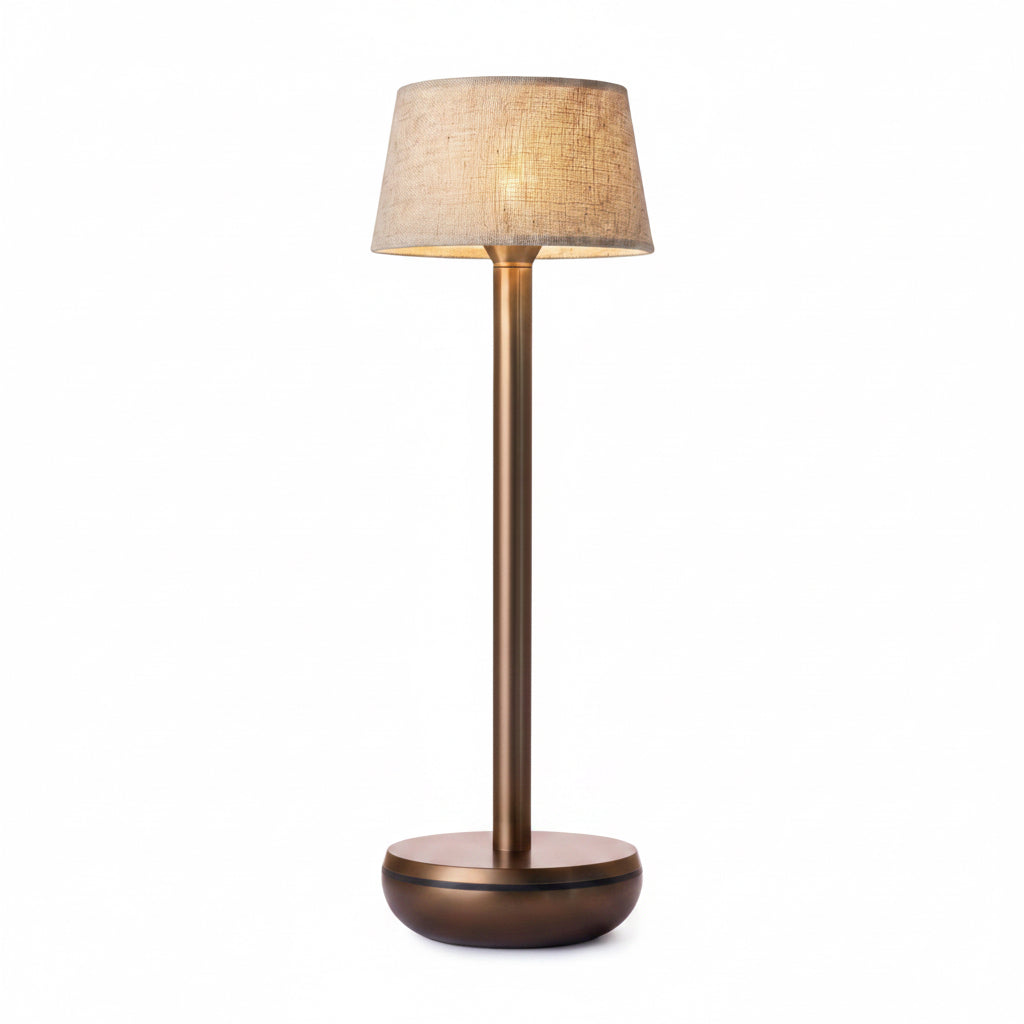 A modern table lamp with a textured beige fabric drum shade, a slim bronze-finished metal stem, and a round bronze base, shown illuminated against a plain white background.