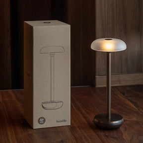 A Bumble Air LED floor lamp with a frosted white dome shade and a dark metallic base, shown next to its brown cardboard packaging box on a dark wood floor.