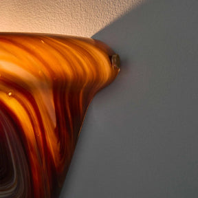 A close-up of a glossy amber and orange marbled glass object against a light gray wall, with warm light reflecting off its curved surface.