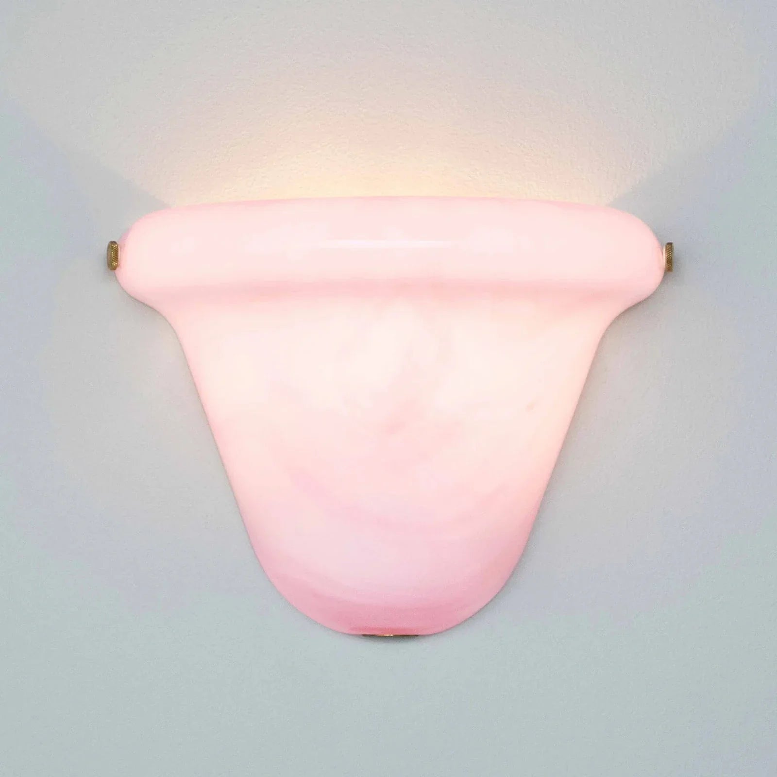 A pink glass wall sconce with a curved, shield-like shade and brass hardware, mounted on a light gray wall and casting a warm glow upwards.