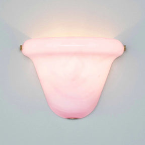 A pink glass wall sconce with a curved, shield-like shade and brass hardware, mounted on a light gray wall and casting a warm glow upwards.