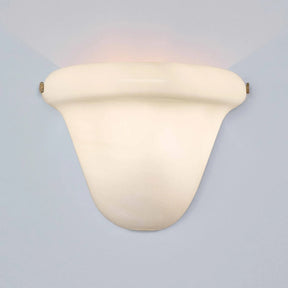 A modern wall-mounted sconce with a curved, shield-like shade in a warm cream or off-white finish, casting a soft warm light upward and downward against a pale blue-gray background.