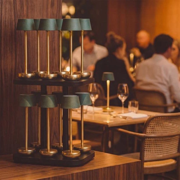 A tiered wooden candle holder with multiple green lampshade-style holders and gold-toned stems, displayed on a wooden table in a warmly lit restaurant setting with blurred diners in the background.