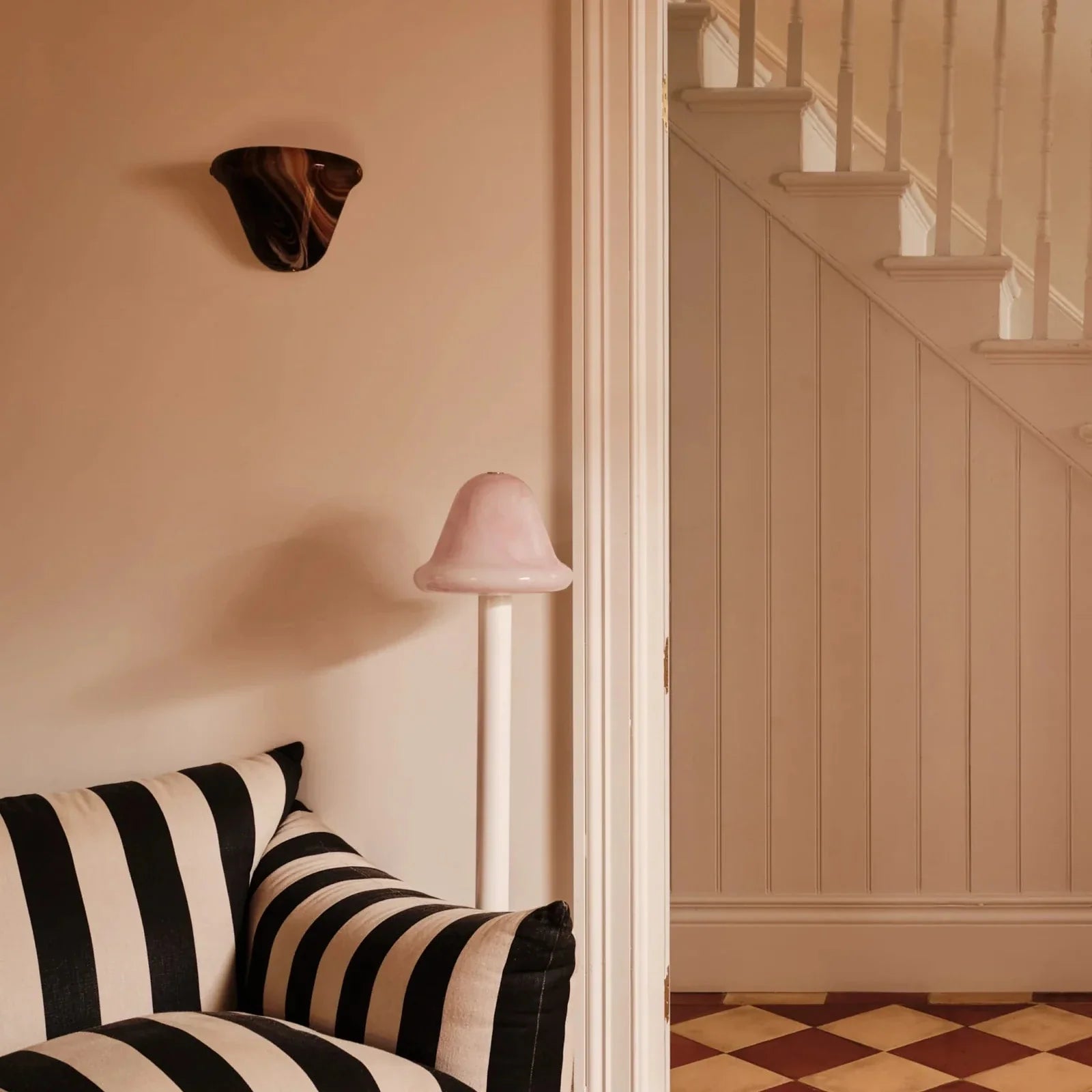 A styled interior corner showing a black and cream striped armchair, a pink floor lamp, a wall sconce, and an open doorway leading to a staircase.