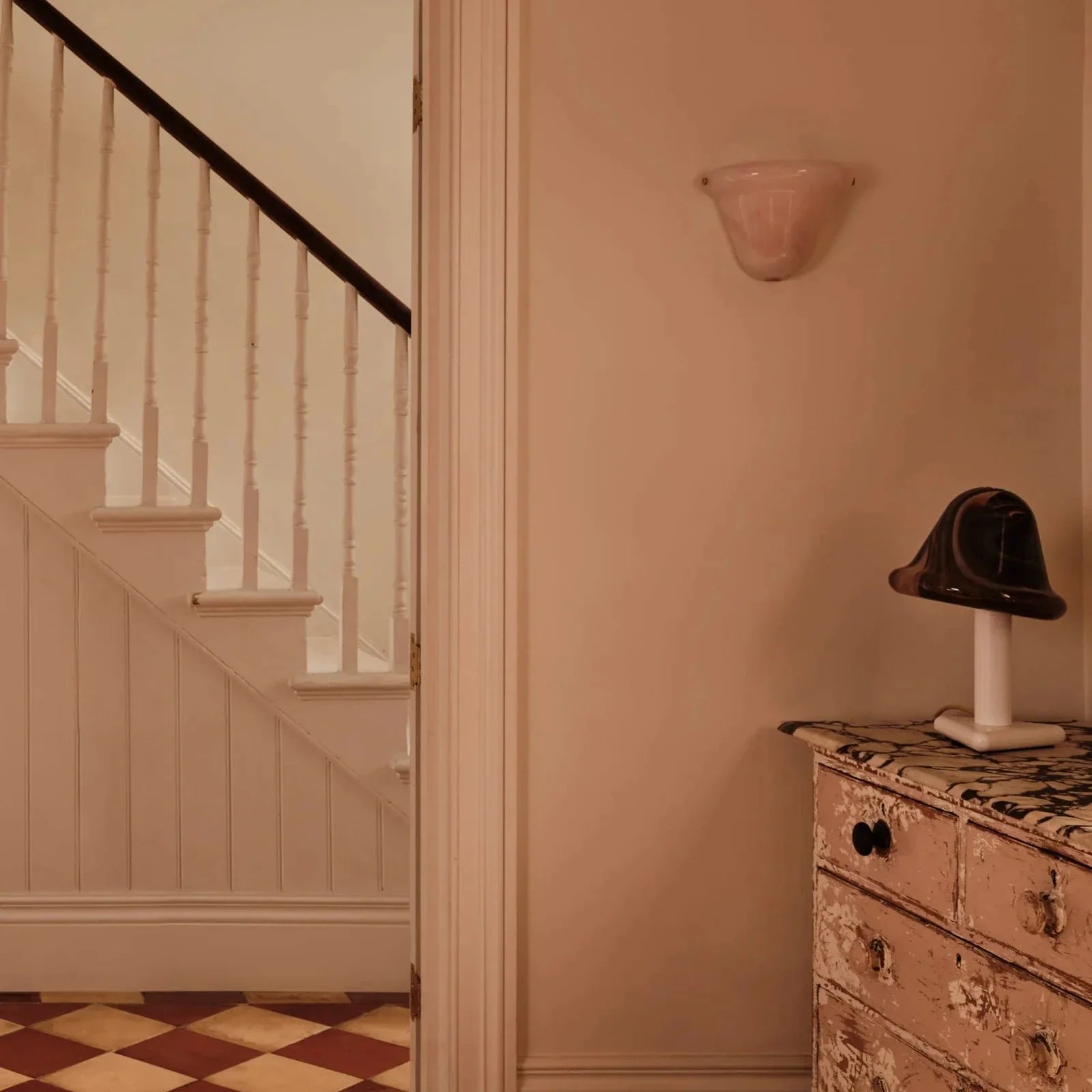 A warm-toned interior hallway scene with a cream-colored wall sconce, a white staircase with a dark handrail on the left, and a distressed pinkish-beige dresser with a small black hat on top in the foreground.
